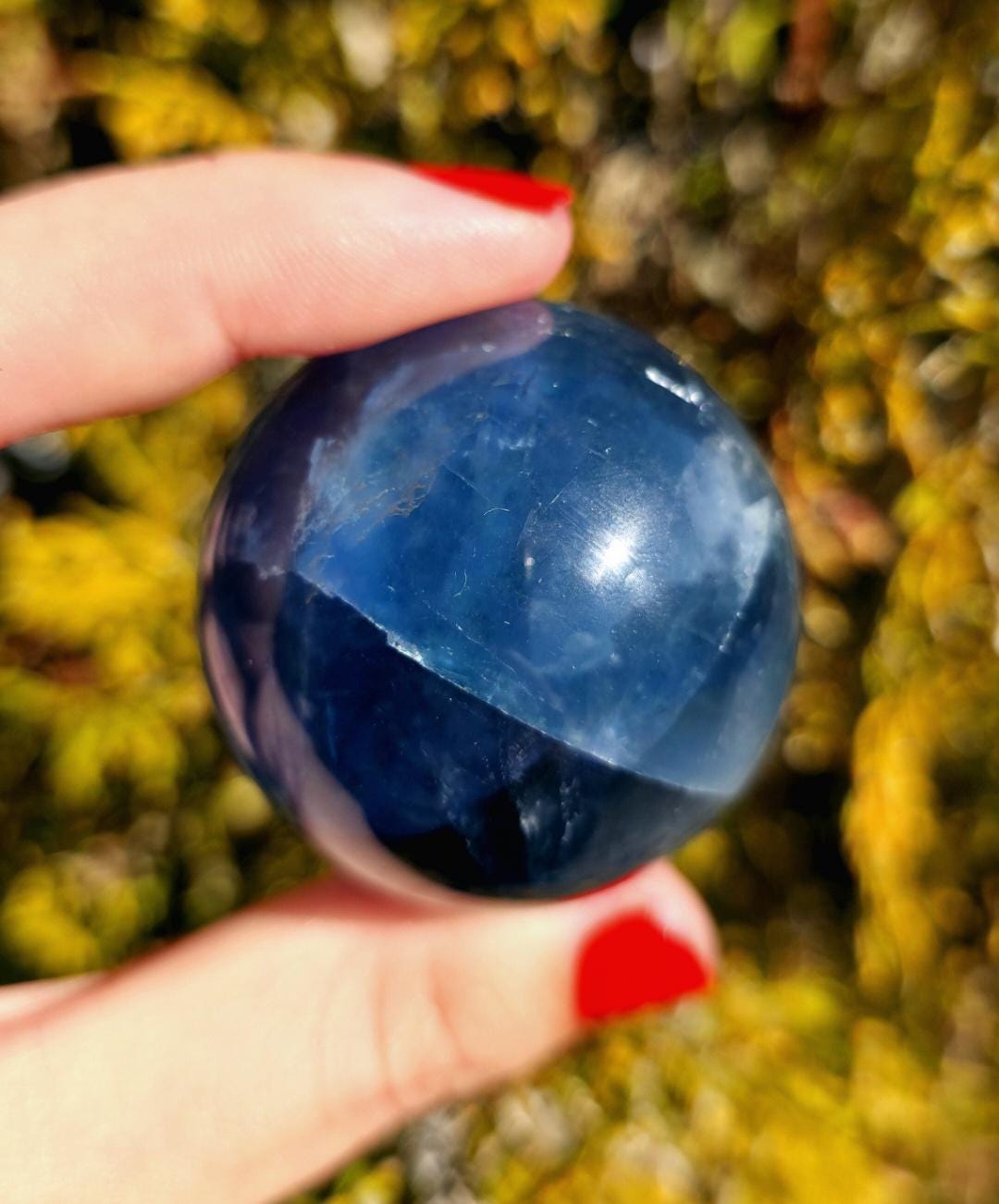 Close up of polished blue fluorite sphere showing natural banding and internal crystal structure, deep blue tones with slight translucency, high polish finish, round fluorite crystal ball, perfect for collectors, crystal display, metaphysical use, and home decor, photographed outdoors in natural light.