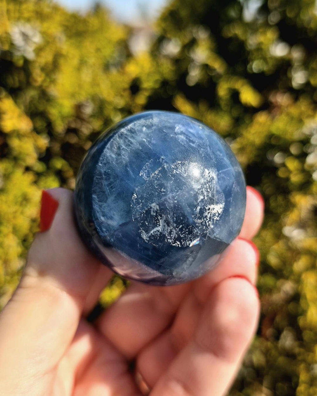 Close up of polished blue fluorite sphere showing natural banding and internal crystal structure, deep blue tones with slight translucency, high polish finish, round fluorite crystal ball, perfect for collectors, crystal display, metaphysical use, and home decor, photographed outdoors in natural light