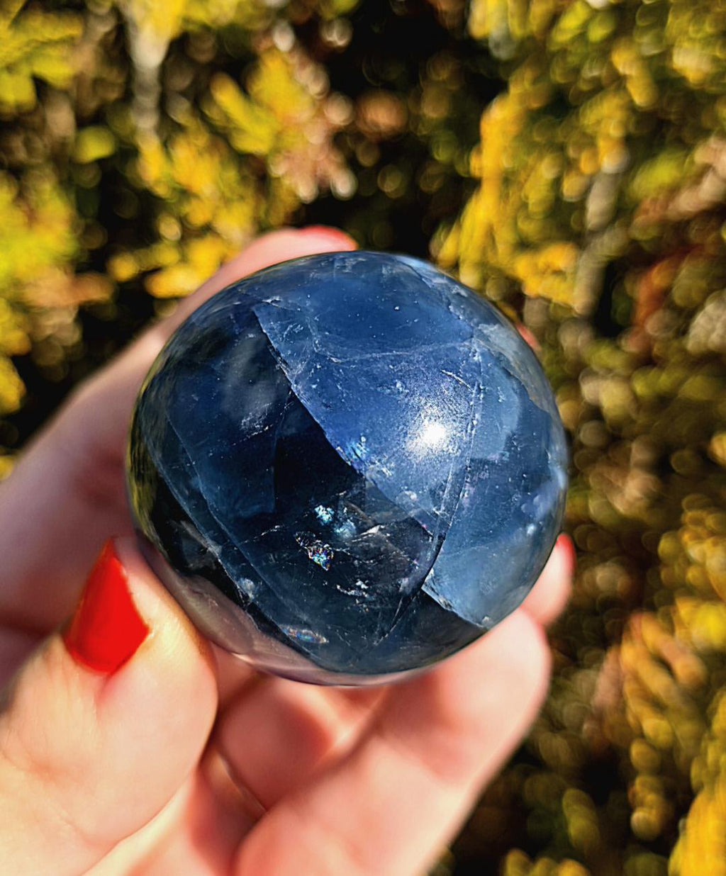 Close up of polished blue fluorite sphere showing natural banding and internal crystal structure, deep blue tones with slight translucency, high polish finish, round fluorite crystal ball, perfect for collectors, crystal display, metaphysical use, and home decor, photographed outdoors in natural light