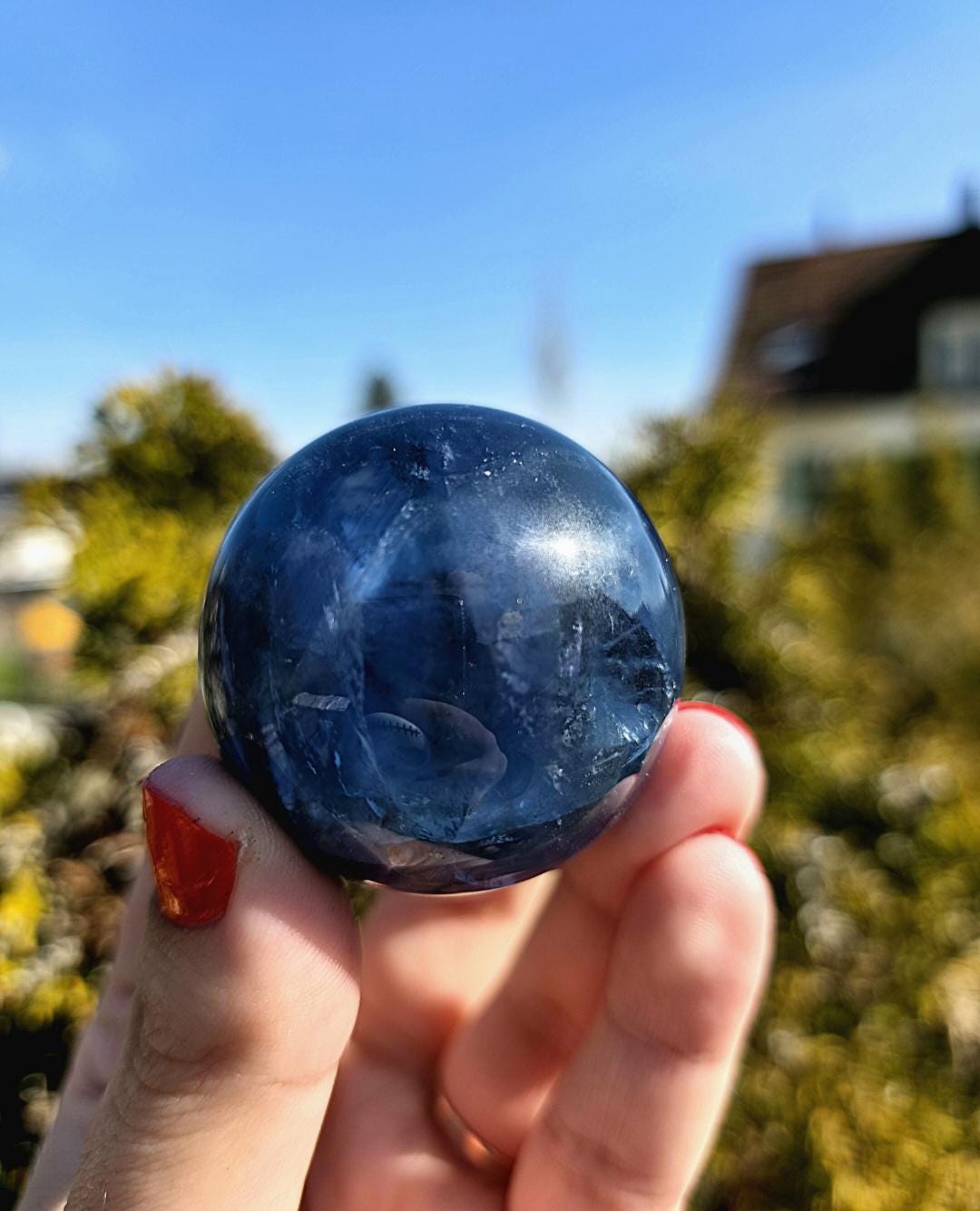 Polished blue fluorite sphere resting in hand against sunlit greenery, showcasing deep blue coloration with natural banding and cloudy inclusions. Bright outdoor lighting enhances clarity and color variation.