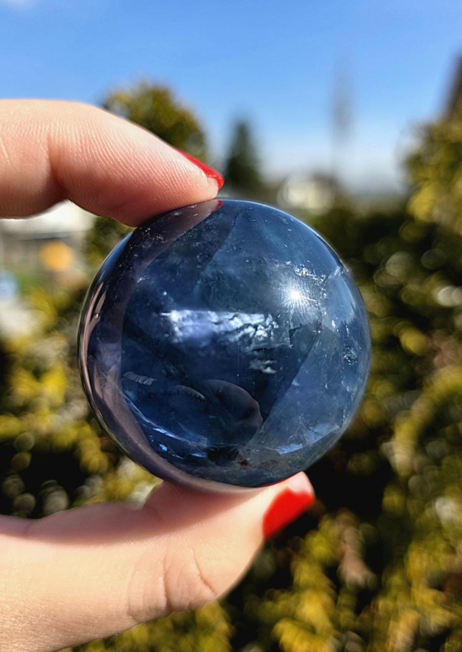 Polished blue fluorite sphere resting in hand against sunlit greenery, showcasing deep blue coloration with natural banding and cloudy inclusions. Bright outdoor lighting enhances clarity and color variation.
