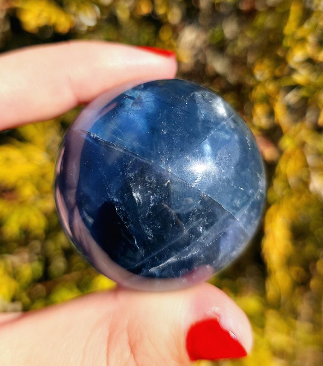 Close up of polished blue fluorite sphere showing natural banding and internal crystal structure, deep blue tones with slight translucency, high polish finish, round fluorite crystal ball, perfect for collectors, crystal display, metaphysical use, and home decor, photographed outdoors in natural light