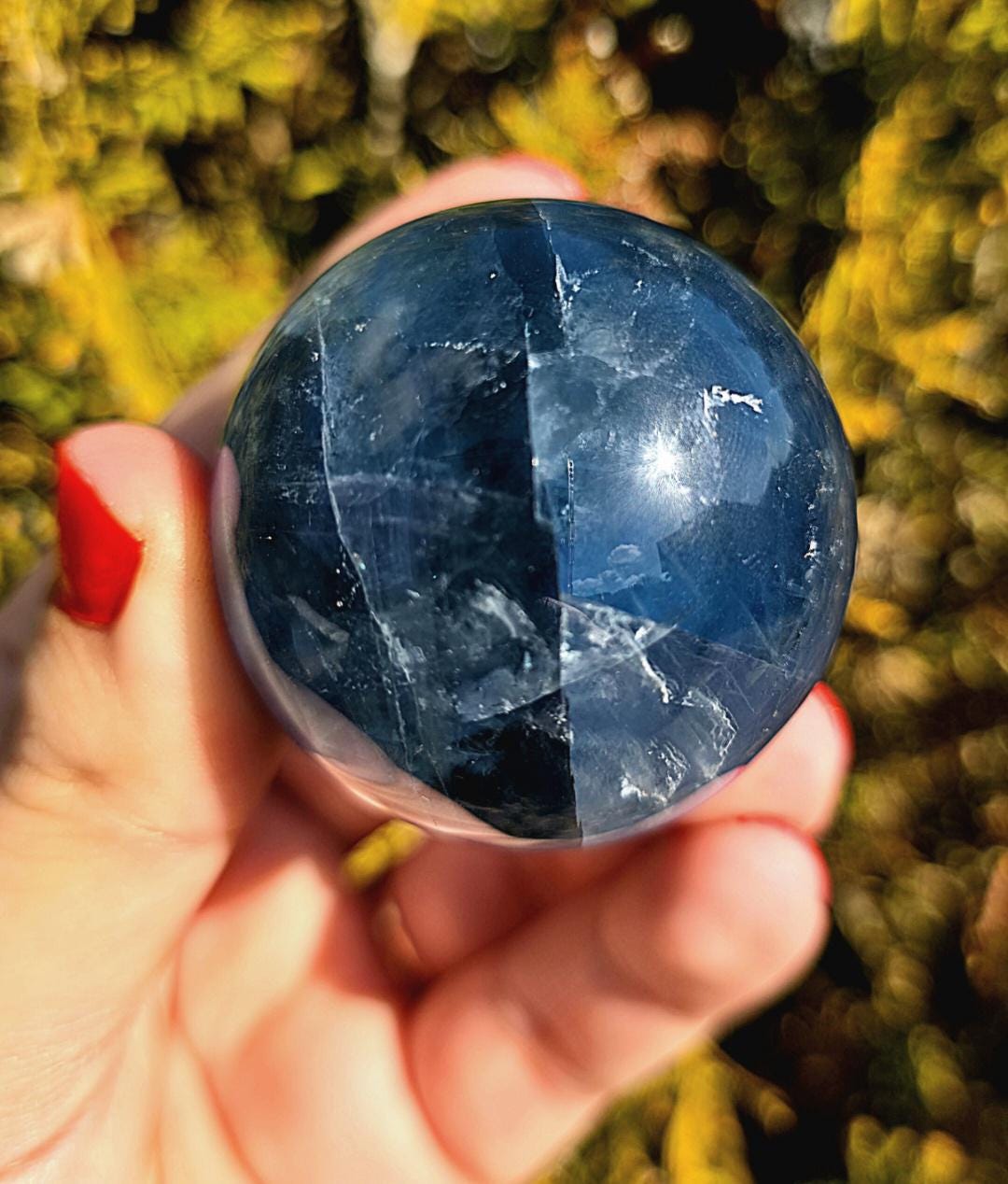 Close up of polished blue fluorite sphere showing natural banding and internal crystal structure, deep blue tones with slight translucency, high polish finish, round fluorite crystal ball, perfect for collectors, crystal display, metaphysical use, and home decor, photographed outdoors in natural light