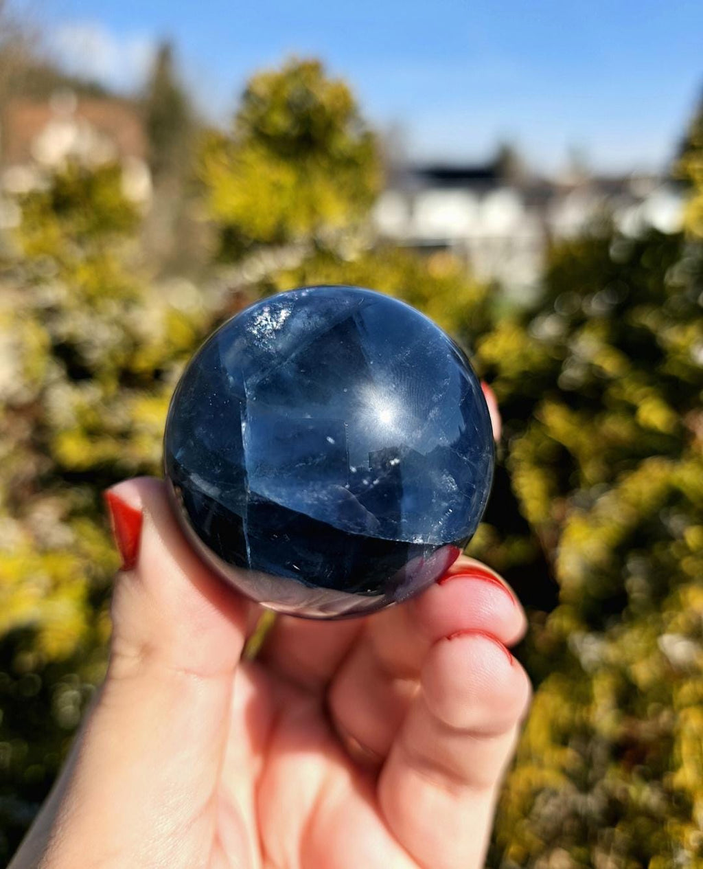 Polished blue fluorite sphere resting in hand against sunlit greenery, showcasing deep blue coloration with natural banding and cloudy inclusions. Bright outdoor lighting enhances clarity and color variation.