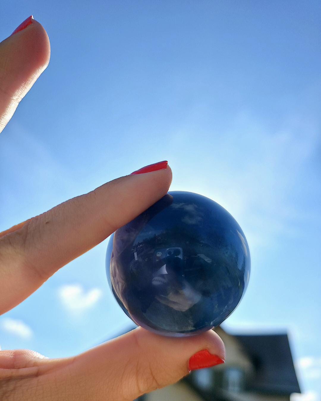 Polished blue fluorite sphere resting in hand against sunlit greenery, showcasing deep blue coloration with natural banding and cloudy inclusions. Bright outdoor lighting enhances clarity and color variation.