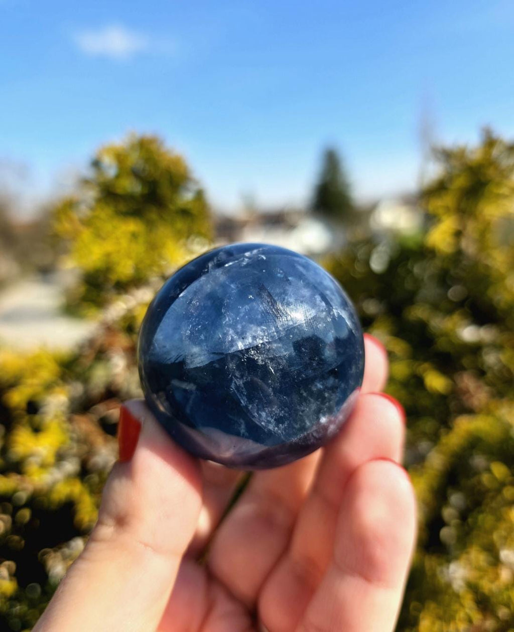 Polished blue fluorite sphere resting in hand against sunlit greenery, showcasing deep blue coloration with natural banding and cloudy inclusions. Bright outdoor lighting enhances clarity and color variation.