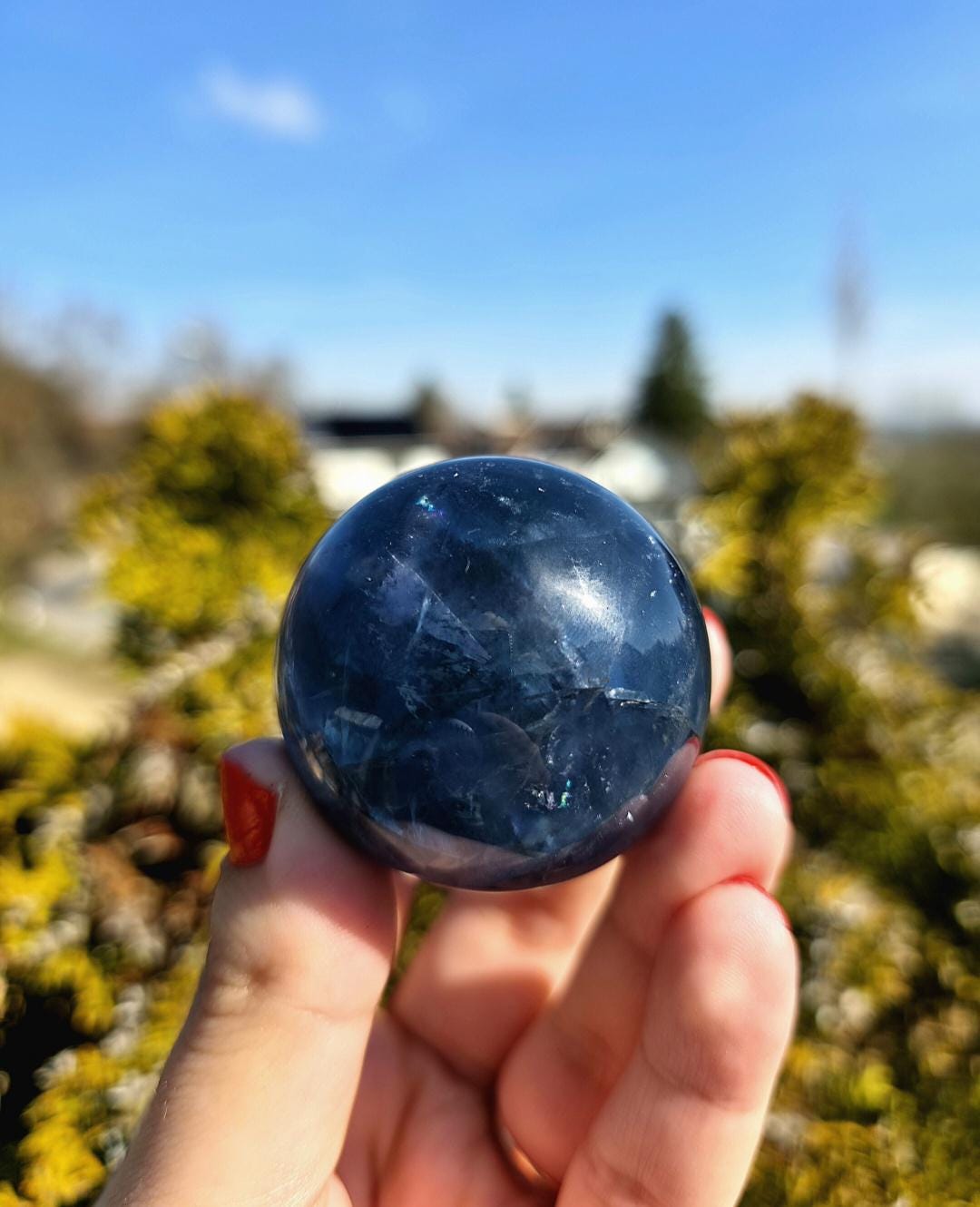 Polished blue fluorite sphere resting in hand against sunlit greenery, showcasing deep blue coloration with natural banding and cloudy inclusions. Bright outdoor lighting enhances clarity and color variation.