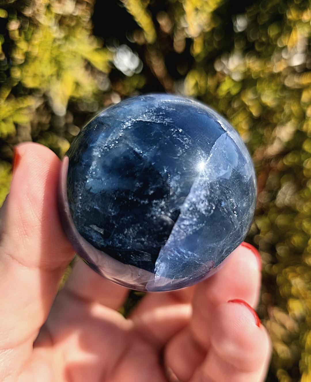 Polished blue fluorite sphere resting in hand against sunlit greenery, showcasing deep blue coloration with natural banding and cloudy inclusions. Bright outdoor lighting enhances clarity and color variation.
