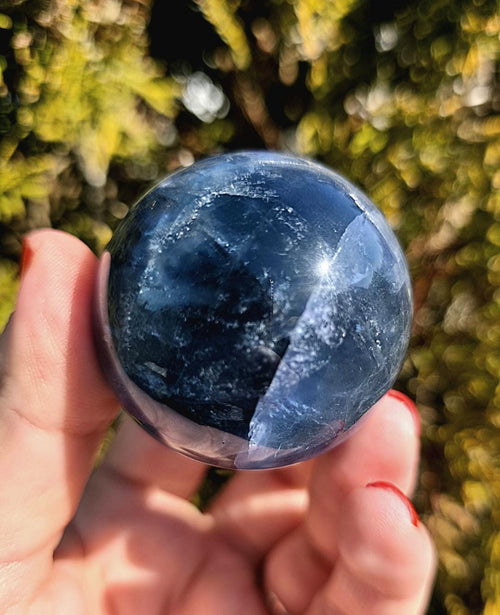 Polished blue fluorite sphere resting in hand against sunlit greenery, showcasing deep blue coloration with natural banding and cloudy inclusions. Bright outdoor lighting enhances clarity and color variation.