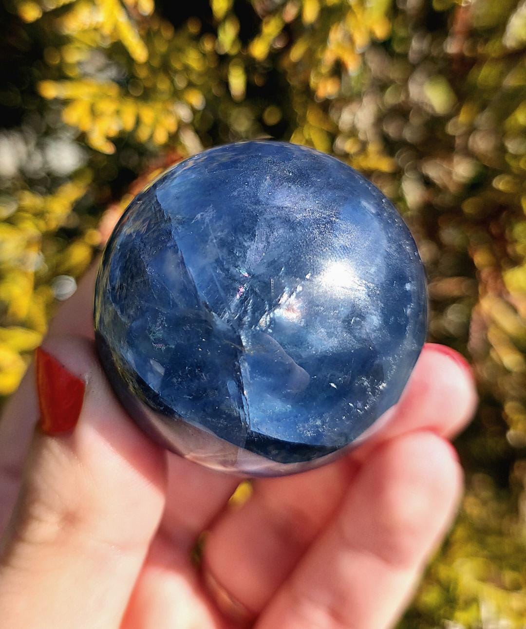 Polished blue fluorite sphere resting in hand against sunlit greenery, showcasing deep blue coloration with natural banding and cloudy inclusions. Bright outdoor lighting enhances clarity and color variation.