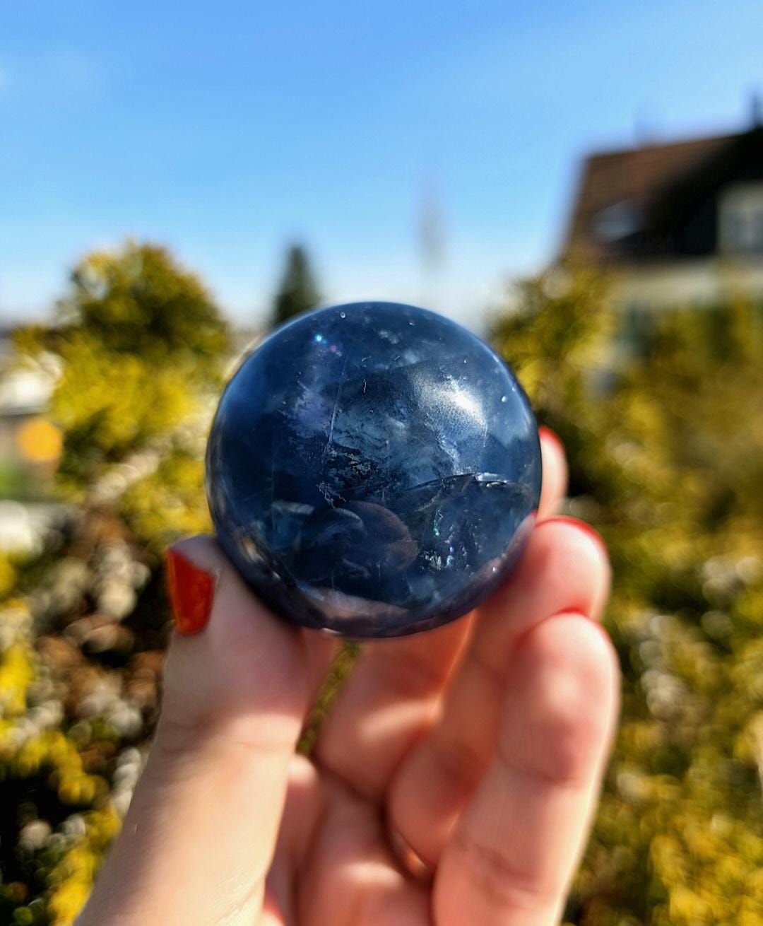Polished blue fluorite sphere resting in hand against sunlit greenery, showcasing deep blue coloration with natural banding and cloudy inclusions. Bright outdoor lighting enhances clarity and color variation.